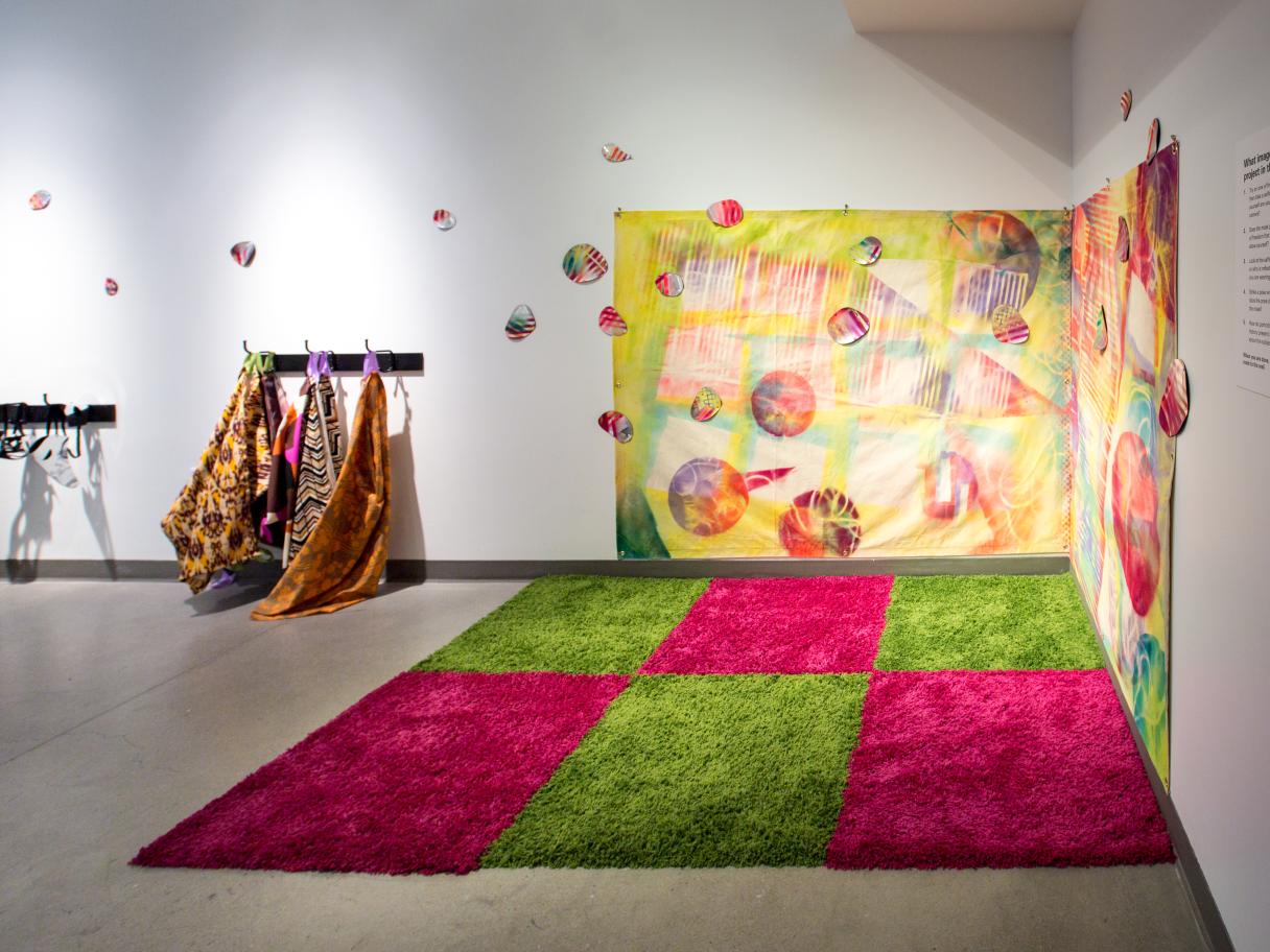 Mirror Masks ACME Lab A green and pink checkered, textured rug sits in the corner of a white room. There's a colorful sheet hanging on the joining walls over the rug. Small colorful circles hang from the ceiling. There are colorful smocks hanging on a coat rack on the wall too.