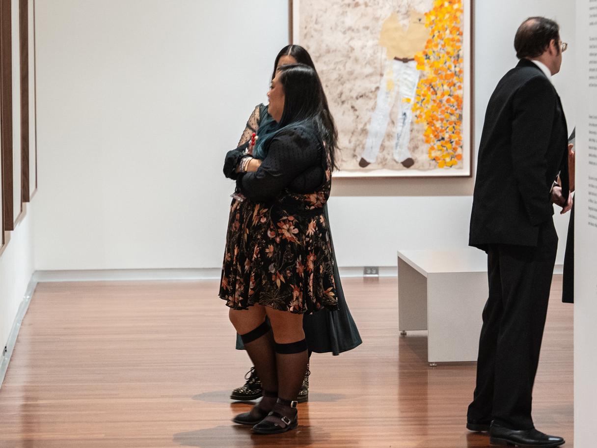 salt 16: Arleene Correa Valencia Two women with long dark hair stand next to one another, speaking and looking at art hung on a wall.
