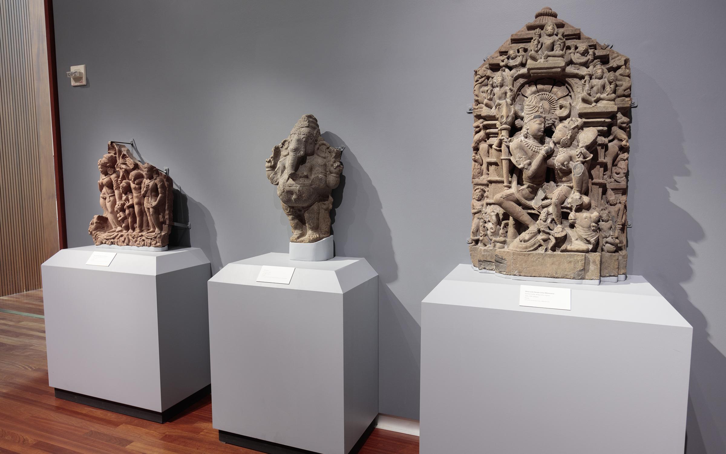 Art of South Asia There are three intricately carved deities lined up on individual stands against a light grey wall.
